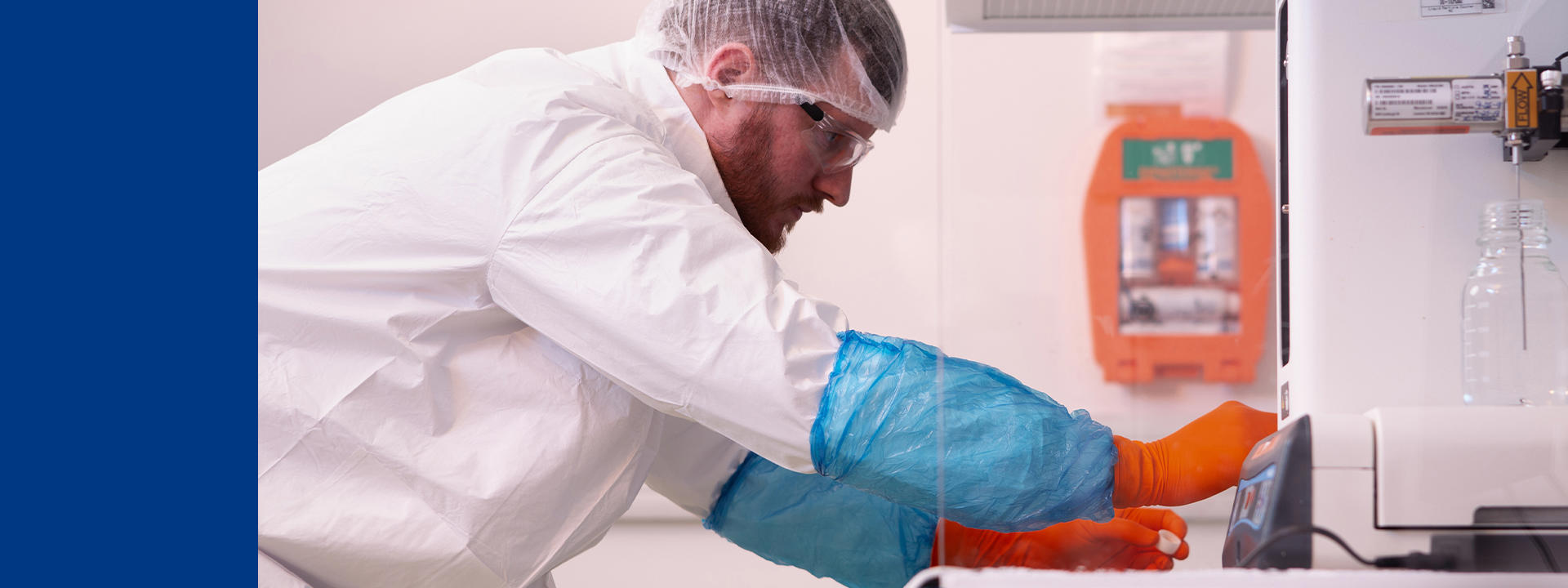 Laboratory professional wearing protective coveralls, gloves, hair cover, and safety glasses, reaching into a laboratory opening or chamber with orange-gloved hands, working in a controlled environment with equipment and safety signage visible in the background.