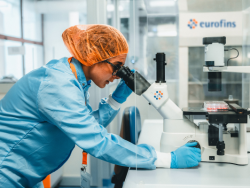 Laboratory professional in cleanroom attire using a microscope to examine a sample on a lab bench, with the Eurofins logo visible on equipment in the background.
