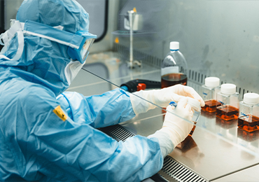 Close-up of a laboratory professional in full protective cleanroom attire, including a blue sterile suit, gloves, and face covering, working inside a controlled environment while handling a tray of small sample containers. Several bottles filled with amber-colored liquid are arranged on the work surface nearby, along with additional lab equipment used for precise sample preparation