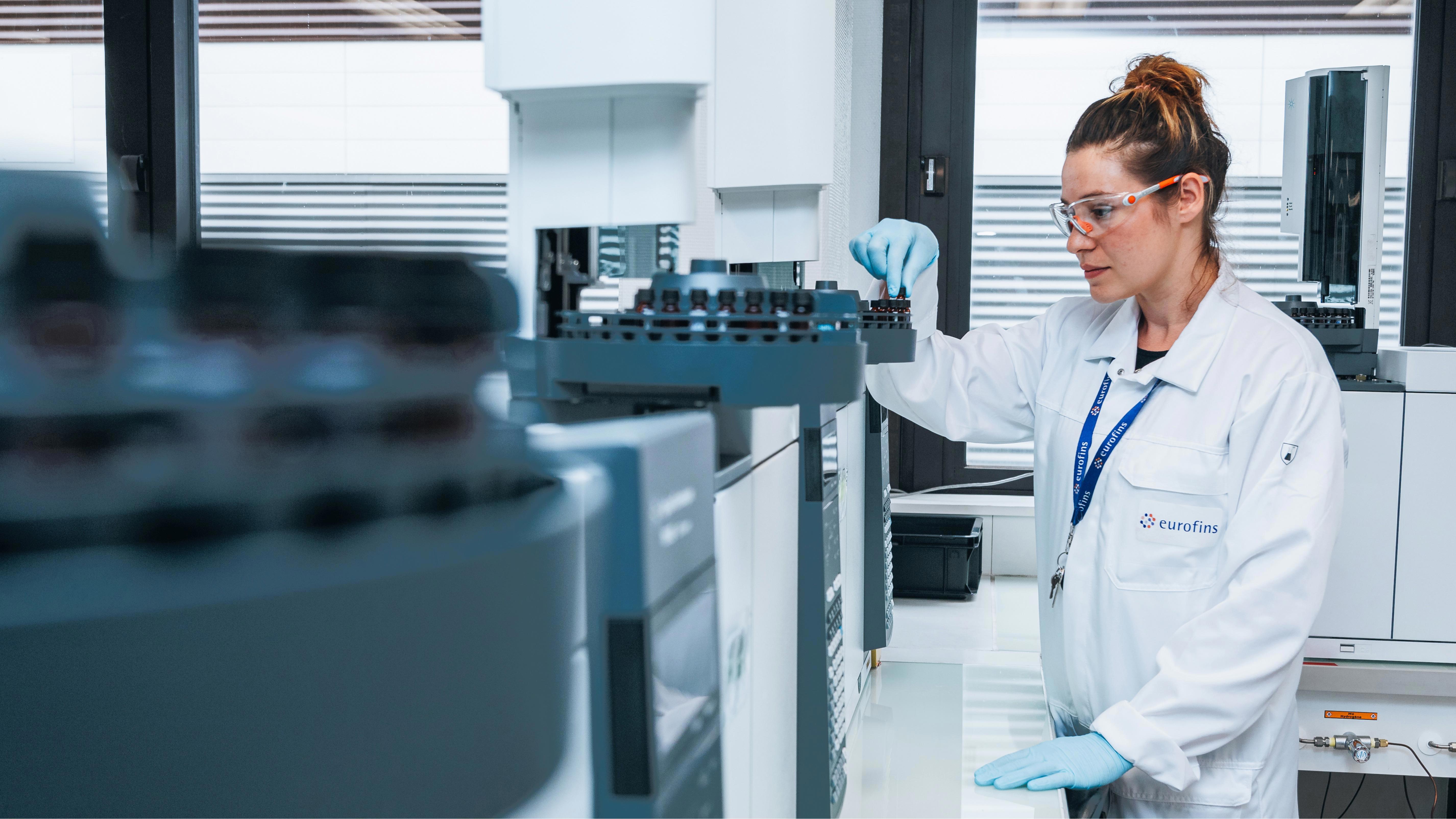 A female laboratory technician placing a vial into a machine to be tested.  The woman is wearing a white lab coat, gloves and safety glasses.