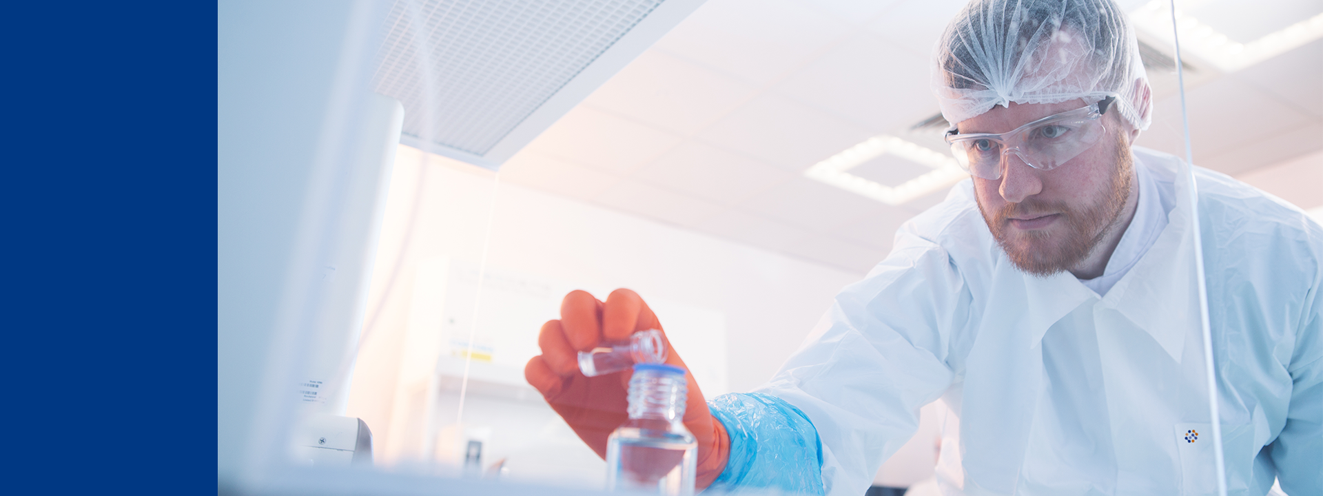 Close-up of a laboratory professional wearing protective gloves, lab coat, hair cover, and safety glasses, carefully holding a small vial of liquid. The individual is viewed through a transparent surface, with a softly lit laboratory environment and equipment visible in the background.