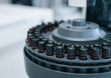 Close-up of a laboratory centrifuge holding multiple small sealed vials arranged in a circular rotor, with dark caps and liquid samples visible, inside a clean analytical lab setting.