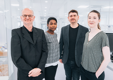Group of four professionals standing together in a bright, modern office or laboratory hallway with glass walls, wearing business and business-casual attire, positioned side by side and facing the camera.