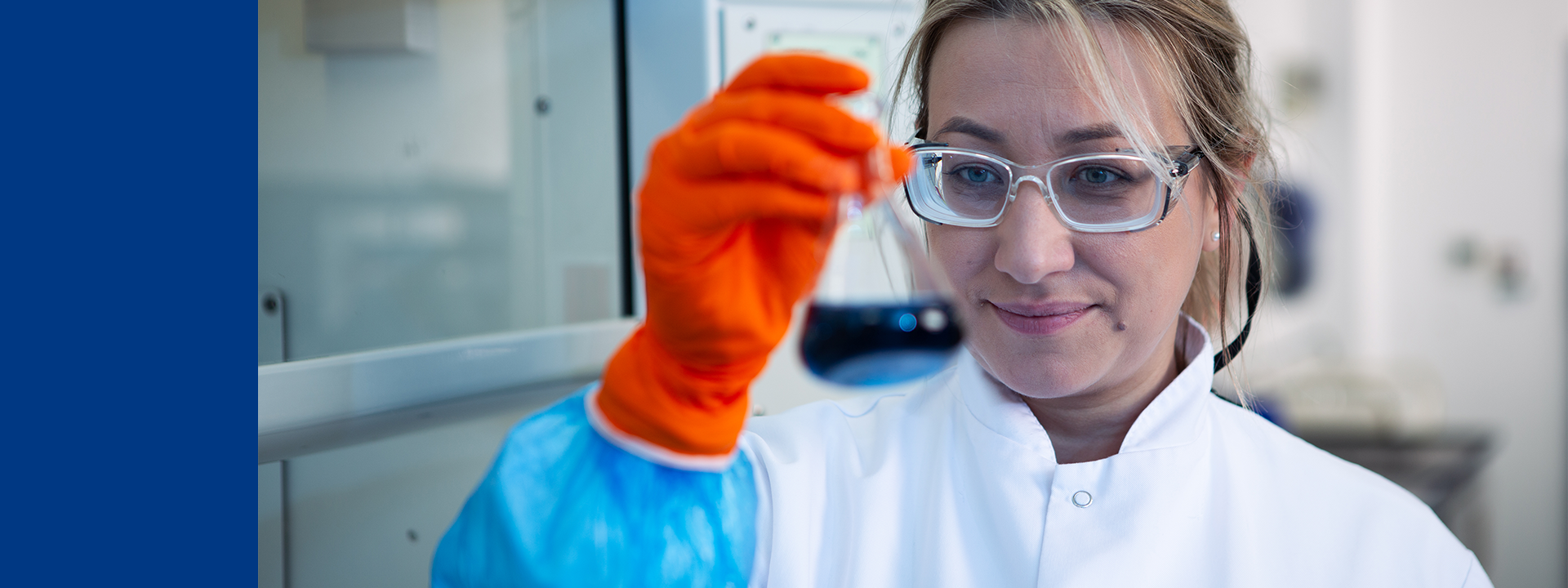 Close-up of a laboratory professional wearing safety glasses and a lab coat, holding a small sample up for inspection while wearing a bright orange protective glove, in a clean lab environment with equipment in the background.