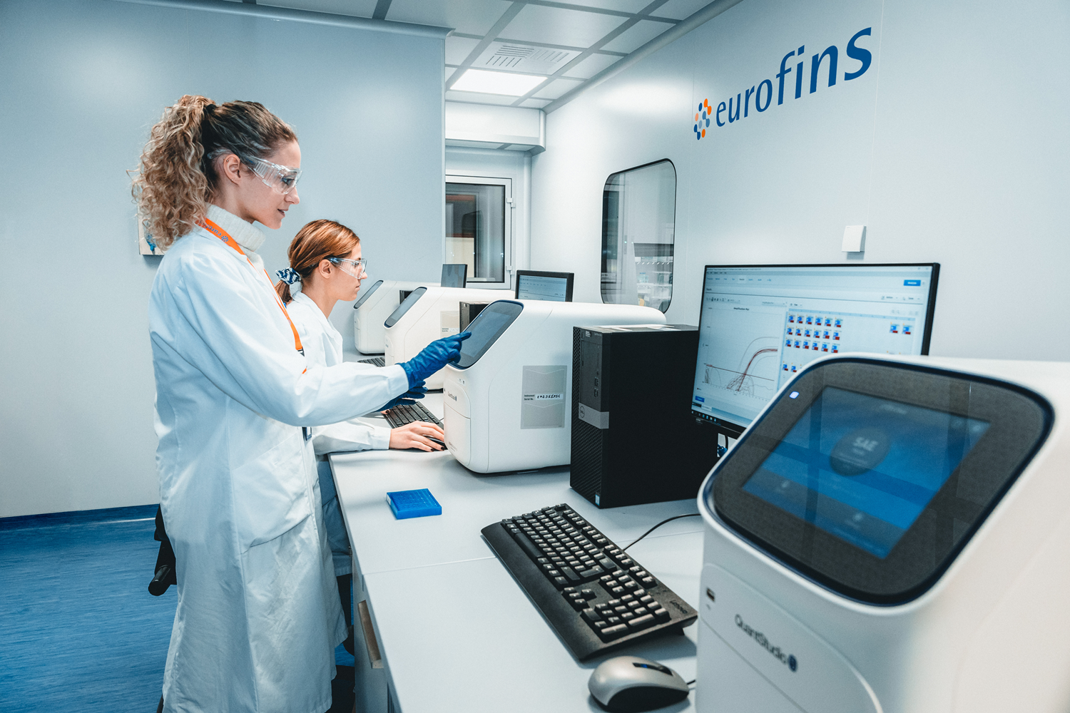 Laboratory professionals in white lab coats working with analytical instruments in a clean lab, with one reviewing a tablet while another operates equipment, and a third working at a nearby station.